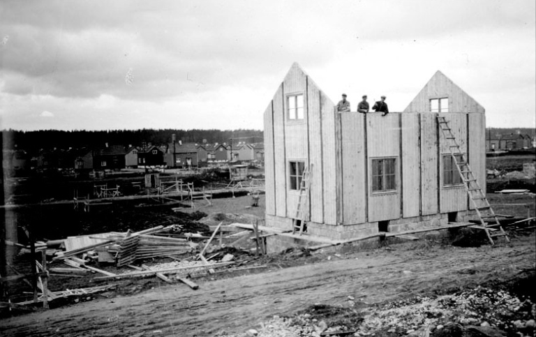 Svartvitt foto av tvåplansvilla i trä under uppbyggnad.