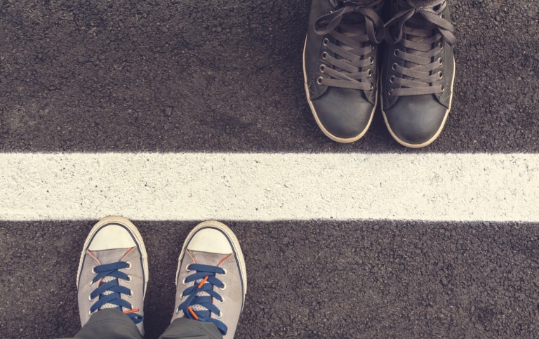 Image of two pairs of feet, spaced apart, with a painted wide line on the tarmac between them.